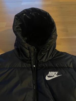 Svart Nike pufferjacka Therma-Fit S - Svart pufferjacka från Nike med Therma-Fit-teknologi. Jackan har huva med dragsko, stor Nike-logga på bröstet och dragkedja framtill. Tillverkad i syntetmaterial som håller värmen under kalla dagar. Perfekt för dig som vill ha en sportig och clean look. Nypris 3200kr