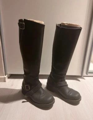 Johnny Bulls höga stövlar - Säljer ett par svarta höga boots i skinn med rund tå och platt sula. De har två silvriga spännen, ett vid ankeln och ett upptill, samt dragkedja på insidan. Perfekta för dig som gillar en edgy stil och vill ha boots som sticker ut. Säljes i fint skick stått oanvända under lång tid nu❣️