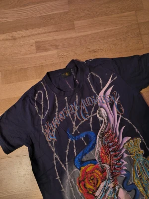 Christian Audigier t-shirt med tryck - Säljer en mörkblå t-shirt från Christian Audigier med fet print och gnistrande paljettdetaljer. Framsidan har motiv med ros, vingar och taggtråd, medan baksidan har stort 1958-tryck och Los Angeles-text. Riktigt unik och edgy design för dig som vill sticka ut.