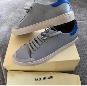 Grå sneakers från Axel Arigato - Snygga grå sneakers från Axel Arigato med blå detalj vid hälen och vit platt sula. Skorna har rund tå och klassisk snörning. Tillverkade i skinn med minimalistisk design och subtil logga på sidan. Perfekta för dig som gillar stilrena och moderna sneakers. Storlek 42. HELT NYA.
