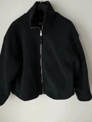 Svart teddyjacka från Monki XS - Mysig svart teddyjacka från Monki i storlek XS. Jackan har hög krage, hel dragkedja framtill och är tillverkad i fluffigt teddy-material. Perfekt för kyliga dagar och ger en cool, avslappnad vibe.