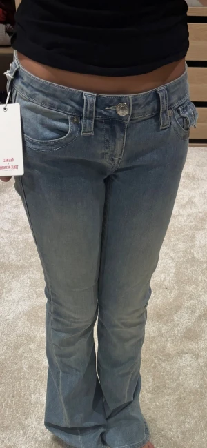 True Religion Joey Low Rise Flare Jeans - Säljer ett par ljusblå Joey Low Rise Flare jeans från True Religion. De har låg midja, utställda ben och klassiska fickdetaljer med knappar bak. Jeansen är i denim med snygga sömmar och coola detaljer som ger en riktigt retro vibe. Helt nya