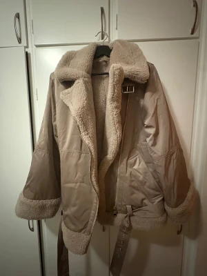 Beige aviator jacka & other stories  - Säljer en beige teddyjacka med fluffig krage och muddar. Jackan har bälte i midjan, dragkedja och stora fickor framtill. Insidan och detaljerna är i mjuk teddy, perfekt för kalla dagar. Oversized passform och väldigt varm! Använd max 5 gånger. Ordinarie pris 1100kr.