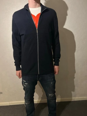 Svart kofta från Selected Homme - Snygg svart kofta från Selected Homme med dragkedja framtill och diskret strukturmönster på bröstet och ärmarna. Modellen har långärmad passform och ribbade muddar vid ärmslut och krage. 