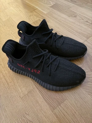 Adidas Yeezy Boost 350 V2 Black/Red - Säljer ett par Adidas Yeezy Boost 350 V2 i svart med röda SPLY-350-detaljer på sidan. Skorna har stickad ovandel, platt sula och snörning. Perfekta för dig som vill ha en clean och hype sneaker med streetkänsla.