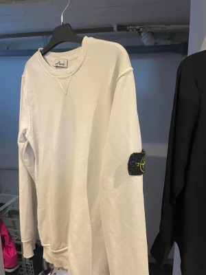 Vit Stone Island sweatshirt - Vit sweatshirt från Stone Island med klassisk rund halsringning och ribbade muddar. Tröjan har den ikoniska svarta logotyp-patchen på vänster ärm och är tillverkad i mjuk bomull. Perfekt för en clean och stilren look. Står XL men den sitter som en M