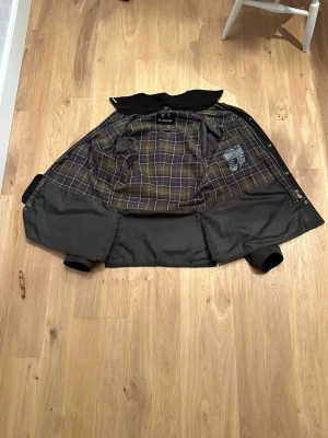 Barbour Ashby dark olive  - Klassisk   vaxad jacka från Barbour med rutigt innerfoder i beige, grått och gult. Jackan har tryckknappar, krage i manchester och två stora fickor framtill. Perfekt för dig som gillar tidlös brittisk stil och funktionella ytterplagg. Inköpt för ca 1 år sedan på NK men ej blivit använd. 