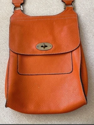 Orange axelväska från Mulberry i skinn - Snygg orange axelväska från Mulberry i grained skinn med klassiskt vridlås i mässing och präglad logga framtill. Väskan har lock, justerbar axelrem och kontrastsömmar som ger en cool touch. Perfekt för dig som vill sticka ut med färg och stil.