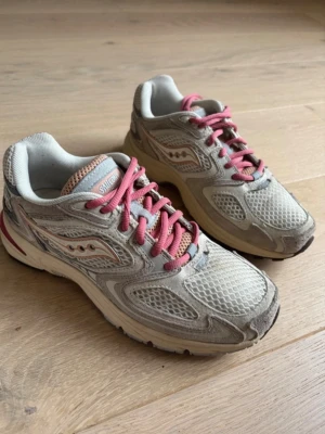Saucony sneakers - Saucony sneakers i ljusgrå mesh och syntet med rosa snörning och detaljer. Skorna har rund tå, platt sula och ventilerande material. Perfekt för dig som gillar sportig och retro stil. Storlek 37.5 