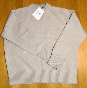 Ljusbrun stickad tröja från Ami - Snygg beige stickad tröja från Ami Alexandre Mattiussi med diskret broderad logga med hjärta på bröstet. Tröjan har ribbade muddar vid hals, ärmslut och nederkant samt en klassisk rund halsringning. Perfekt för dig som gillar stilrena och mjuka plagg.