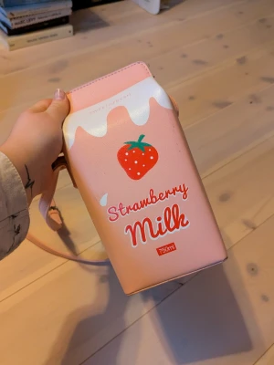 Rosa Strawberry Milk axelväska - Supergullig axelväska formad som en jordgubbsmjölk-förpackning. Väskan är ljusrosa med vita detaljer och har ett tryck av en jordgubbe och texten 'Strawberry Milk'. Tillverkad i syntetmaterial och har justerbar axelrem. Perfekt för dig som gillar unika accessoarer!