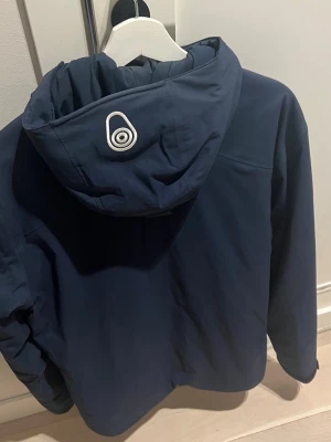 Mörkblå Sail Racing Gore-Tex skidjacka - Mörkblå jacka från Sail Racing med Gore-Tex-material, huva och två vertikala bröstfickor med dragkedja. Jackan har logga på bröstet och ärmen samt justerbar huva. Perfekt för dig som vill ha en snygg och funktionell jacka med sportig vibe. **Äkta** 