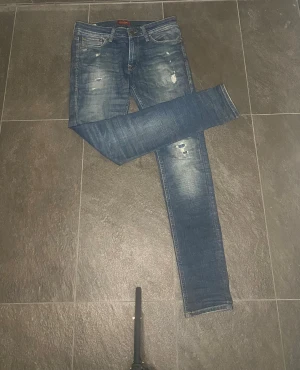 Jack & Jones jeans blå  - Snygga Replay Anbass jeans i blå tvätt med slitningar och skinny passform. Klassisk femficksmodell med läderpatch bak i midjan. Jeansen har smal siluett och är tillverkade i stretchigt denim för skön komfort.