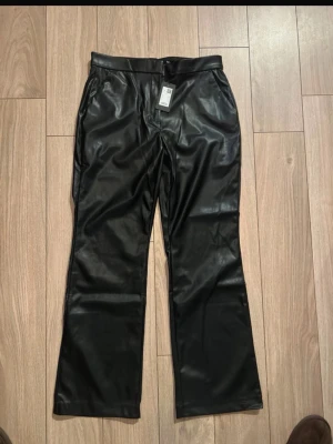 Svarta raka byxor i skinnimitation - Snygga svarta byxor i skinnimitation med raka ben och klassisk midja. Byxorna har sidfickor fram och passpoalfickor bak, samt stängs med knapp och dragkedja. Perfekta för en edgy look.