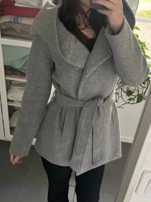 Grå kappa med huva och bälte - Säljer en snygg grå kappa med stor huva och knytskärp i midjan. Jackan har dubbelknäppning framtill, markerad midja och långa ärmar. Den har super fin passform och är strukturerad.
