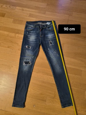 Blå Catem's  dsquared2 Jeans med slitningar - Säljer ett par blå Catem's dsquared2 Jeans med coola slitningar och patchade hål på framsidan. Jeansen har skinny passform, fem fickor och detaljer som färgstänk och en röd läderpatch bak. Perfekt för dig som gillar en edgy streetstil.