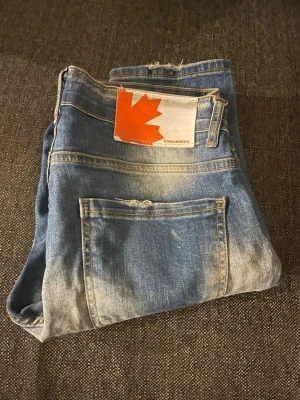 Dsquared2 blå slitna jeans, storlek 32 - Riktigt snygga blå jeans från Dsquared2 med slitningar, hål och färgstänk för en cool, edgy look. Klassisk femficksmodell med knappgylf och tydlig Dsquared2-logga. Passar dig som gillar streetstyle och vill sticka ut.