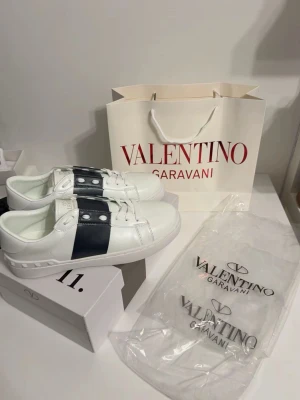 Valentino Garavani Open sneakers vit/blå - Snygga Valentino Garavani Open sneakers i vitt skinn med bred marinblå läderdetalj och silverfärgade nitar på sidan. Klassisk rund tå, platt sula med mönstrad undersida och vita snören. Perfekt för dig som gillar exklusiv streetstyle.