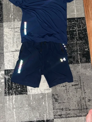 Blå träningsshorts från Under Armour - Snygga marinblå träningsshorts från Under Armour med regnbågsfärgat logotryck och text på benet. Shortsen har elastisk midja med snörning och är gjorda i ett lätt och snabbtorkande syntetmaterial. Perfekta för gymmet eller löprundan.