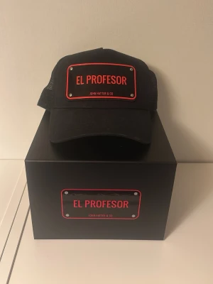Svart keps John Hatter & Co El Profesor - Svart keps från John Hatter & Co med röd och svart metallskylt framtill där det står 'El Profesor'. Kepsen har mesh på sidorna och justerbar snapback baktill. Levereras med originalförpackning och dustbag.