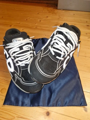 Svarta sneakers med vita detaljer - Säljer ett par svarta sneakers med vita kontrastdetaljer och snörning. Skorna har en chunky platt sula och ett stort på sidan. Materialet är en mix av mesh och syntet, vilket ger en sportig och modern vibe. Perfekt för dig som gillar exklusiva streetwear.