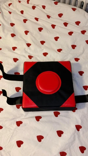 Svart och röd boxningsdyna med kardborre - Boxningsdyna i fyrkantig form med svart och röd design, rund röd mitt för slagträff. Dynan har breda kardborreband för enkel montering på vägg. Materialet är slitstark syntet med vadderad mitt för extra komfort vid träning.