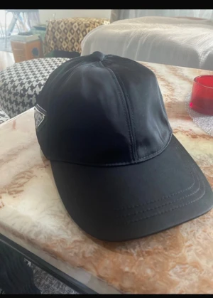 Svart keps från Prada i nylon - Snygg svart keps från Prada med klassisk böjd skärm och ikonisk metallogo på sidan. Tillverkad i glansigt nylonmaterial som ger en lyxig känsla. Perfekt accessoar för att lyfta din outfit med en exklusiv touch.