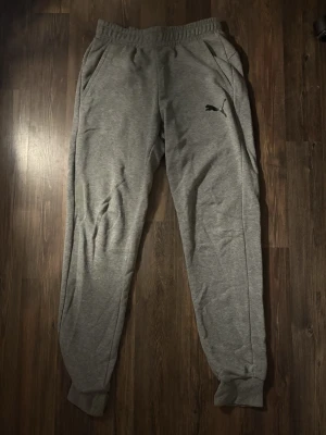 Grå mjukisbyxor från Puma - Snygga grå mjukisbyxor från Puma med svart logga på vänster ben. Byxorna har elastisk midja, ribbade muddar vid bensluten och två sidofickor. Perfekta för chill eller träning, tillverkade i mjukt bomullsmaterial för maximal komfort.