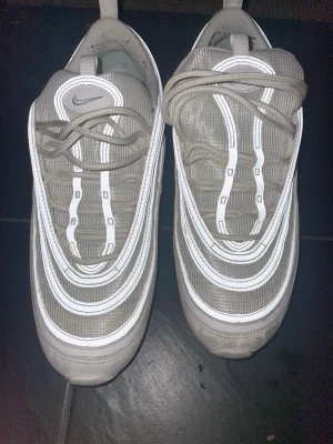 Nike Air Max 97 i beige och vitt - Nike Air Max 97 sneakers i beige och vitt med reflekterande detaljer. Klassisk vågig design, snörning och rund tå. Ovandel i syntet och textil, sula i gummi. Perfekta för dig som gillar en clean och modern look.