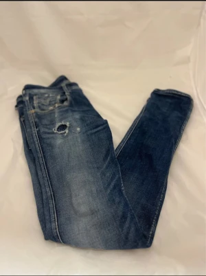 Replay Anbass jeans blå herr - Snygga Replay Anbass jeans i klassisk blå tvätt med slitningar och patchad håldetalj framtill. Jeansen har fem fickor och bruna läderpatch bak. Tuffa kontrastsömmar och röd Replay-logga på bakfickan. Ingen slitning syns in till huden 👍🏽