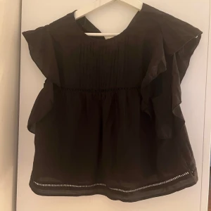 Svart blus med volangdetaljer - Snygg svart blus med volangdetaljer längs axlarna och ärmlös design. Blusen har rund halsringning, fina veck framtill och dekorativa hålbroderier vid midjan och nederkanten. Perfekt för dig som gillar en feminin och trendig look. Från hm i storlek S, Aldrig använd💕