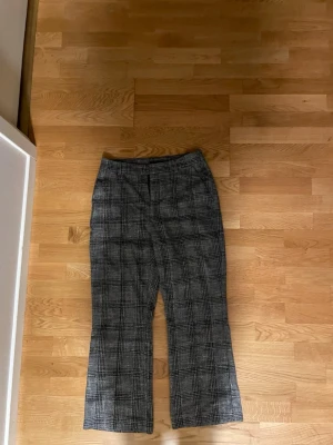 Rutmönstrade kostymbyxor från Lindex - Snygga gråsvarta kostymbyxor från Lindex i storlek 38. Byxorna har ett klassiskt rutmönster, raka ben och midjehällor för bälte. Perfekta för en stilren look och passar till många olika outfits.