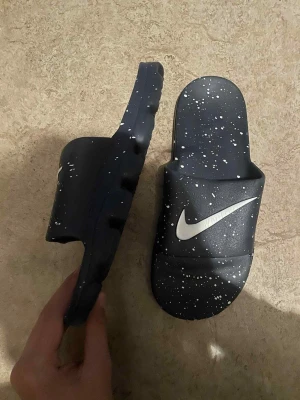 Svarta Nike sandaler med vita stänk - Svarta Nike sandaler med coolt vitt stänkmönster och stor vit Swoosh-logga. Gjorda i syntetmaterial med en chunky, mönstrad sula för extra komfort. Perfekta för sommaren och chill dagar. Snygg och modern look som sticker ut.