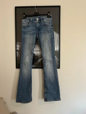 Blå bootcut jeans  - Snygga blå lågmidjade jeans med klassisk bootcut passform och ljus tvätt. Modellen har fem fickor, silverfärgade knappar och bälteshällor. Jeansen är tillverkade i jeansmaterial och har en cool, lite sliten look vid bensluten.
