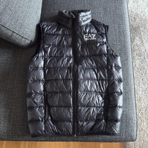 Svart dunväst från EA7 Emporio Armani - Snygg svart dunväst från EA7 Emporio Armani med quiltad design och hög krage.  Perfekt för lager-på-lager och ger en sportig vibe. Materialet är glansigt och lätt, vilket gör den enkel att bära. Inga defekter utan är nästan i nyskick. Säljer då den är för liten för mig