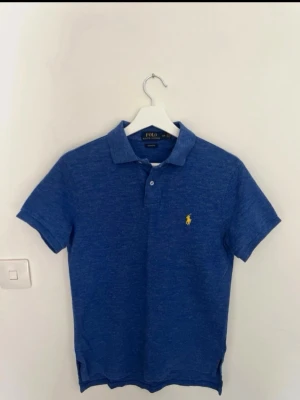 Blå pikétröja från Polo Ralph Lauren - Snygg blå pikétröja från Polo Ralph Lauren med klassisk krage och två knappar framtill. Tröjan har det ikoniska gula Polo-logot på bröstet och är tillverkad i mjuk bomull. Passformen är custom fit och den har korta ärmar. Perfekt för en chill och stilren look.