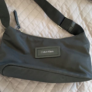 Svart axelväska från Calvin Klein - Snygg svart axelväska från Calvin Klein med justerbar rem och tydlig logga framtill. Väskan har en praktisk dragkedja med CK-detalj och insida med logotyp. Materialet är slitstark polyester och detaljer i skinn. Perfekt storlek för det viktigaste.
