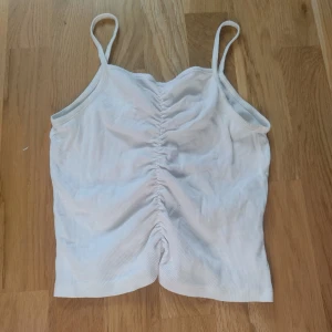 Vit ribbad linnetopp från Kappahl - Säljer en vit ribbad linnetopp från Kappahl med smala axelband och snygg rynkdetalj framtill. Toppen är croppad och har en enkel, fräsch look som passar perfekt till sommaren. Materialet är mjukt och stretchigt för en bekväm känsla.