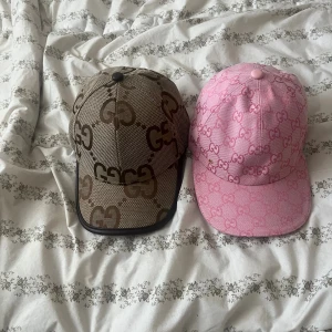 Gucci kepsar med GG-mönster - Två snygga Gucci kepsar med klassiskt GG-mönster. Ena kepsen är beige och brun med stora GG-loggor, den andra är rosa med rosa GG-loggor. Båda har justerbar rem bak och är tillverkade i bomull. Perfekt för dig som vill ha en lyxig och trendig accessoar.