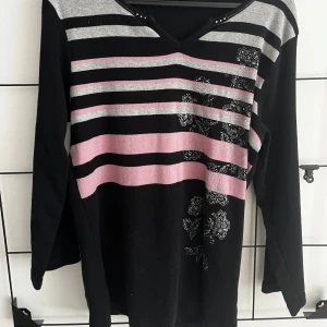 Randig svart tröja med blommor - Svart långärmad tröja med v-ringning och små nitar vid halsen. Framsidan har grå och rosa ränder samt blommiga detaljer i silver. Tröjan har en mjuk känsla och är perfekt för dig som gillar en stilren men ändå lekfull look. Köpt på Vinted. Storlek M