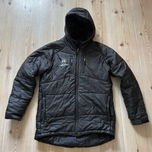 Svart pufferjacka från Unihoc - Svart pufferjacka från Unihoc med huva och dragkedja framtill. Jackan har två sidofickor, en bröstficka med dragkedja och logga på bröstet och ryggen. Tillverkad i syntetmaterial och har lång ärm. Perfekt för kyliga dagar.