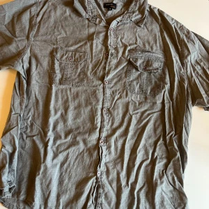 Grå kortärmad skjorta Cosmos 3XL - Grå skjorta från Cosmos i storlek 3XL med korta ärmar och två bröstfickor med knappar. Skjortan har klassisk krage och knäppning framtill. Tillverkad i ett lätt bomullstyg som känns skönt mot huden. Perfekt för dig som gillar en enkel och stilren look.