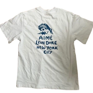 Vit Aimé Leon Dore t-shirt NYC print - Vit t-shirt från Aimé Leon Dore med blått tryck fram och stort print på ryggen med texten 'Aimé Leon Dore New York City' och en grafisk illustration. Klassisk passform och rund hals. Perfekt för dig som vill ha en avslappnad stil med snygga prints. Passar S-M