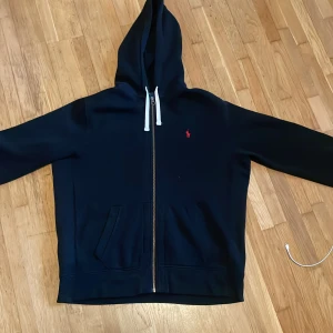 Svart hoodie från Polo Ralph Lauren XL - Svart hoodie från Polo Ralph Lauren i storlek XL. Klassisk design med vit snörning i huvan, dragkedja framtill och röd broderad logga på bröstet. Mjuk bomullskänsla och känguruficka framtill. Perfekt för chill dagar.