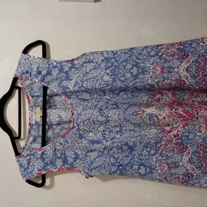 Blommig blå ärmlös klänning Stories - Superfin ärmlös blusar från Stories i blått med vitt och färgglatt blommönster. Klänningen har rund halsringning med kontrastkant i rött och rosa, och ett flowigt fall. Perfekt för dig som gillar boho-vibe och mönster!