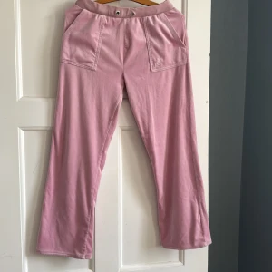 Rosa velourbyxor från Juicy Couture - Mysiga rosa mjukisbyxor från Juicy Couture i velour med bred resår i midjan, stora fickor fram och bak samt broderad JC-logga på ena fickan. Perfekta för en chill och trendig look.