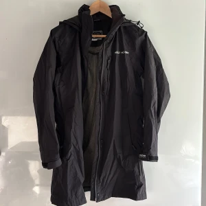 Helly Hansen - Svart regn/vindjacka (dam) - Svart regn/vindjacka (dam) från Helly Hansen med huva och dragkedja framtill. Jackan har meshfoder, praktiska fickor med dragkedja och justerbara ärmslut. Perfekt för blöta dagar och har en stilren, längre passform med logga på bröstet och ryggen.