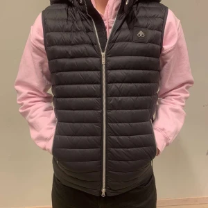 Svart dunväst från Moose Knuckles - Svart quiltad dunväst från Moose Knuckles med huva och dragkedja framtill. Västen har ett diskret logomärke på bröstet och horisontella sömmar som ger en sportig look. Perfekt att slänga över en hoodie för extra värme.