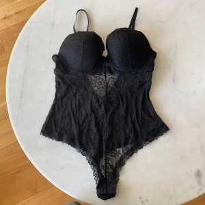 Svart spetsig korsettbody med bygel - Säljer en svart korsettbody med vadderade kupor och bygel, justerbara axelband och snygga spetsdetaljer framtill och baktill. Bodyn har knäppning i grenen och transparent mesh i sidorna för extra stilpoäng.