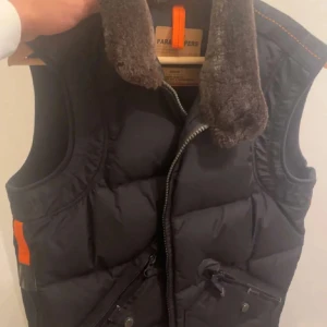 Svart dunväst från Parajumpers - Svart dunväst från Parajumpers med fluffig krage i pälsimitation och orange detaljer. Västen har snedställda fickor med dragkedja och tryckknappar samt en kraftig dragkedja framtill. Perfekt för lager-på-lager under kyliga dagar.